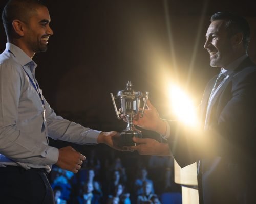 Side view of Caucasian businessman giving trophy to mixed race business male executive on stage in auditorium